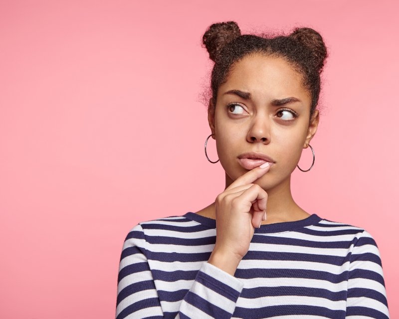 a woman in front of a pink background holds a hand to her chin pondering